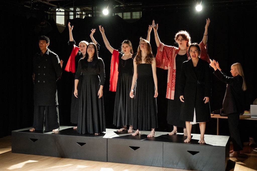 Group of actors wearing bright red or black stand on black risers with their arms raised above their heads in mid-performance during the final presentation of The Winter's Tale.